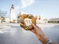 Ręka trzyma obwarzanka krakowskiego na tle Sukiennic i wieży Ratuszowej.