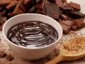 Miska pełna gładkiego, błyszczącego drip czekoladowego, otoczona kawałkami czekolady i drewnianą łyżką.