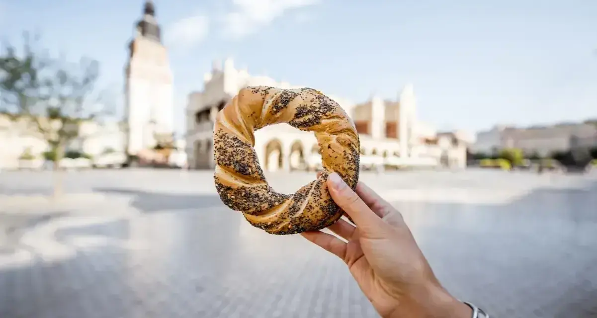 Ręka trzyma obwarzanka krakowskiego na tle Sukiennic i wieży Ratuszowej.