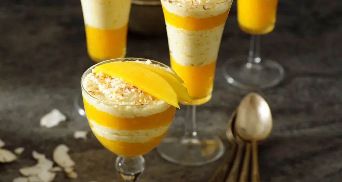 Warstwowy deser z mango, przełożony kremem kokosowym, ozdobiony plasterkiem mango i wiórkami kokosowymi.