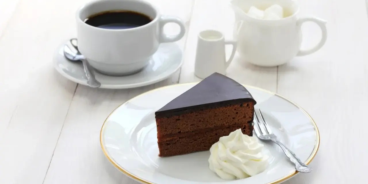 Pyszny tort Sachera z bitą śmietaną i kawą na białym drewnianym stole. Idealny deser na chwilę relaksu.