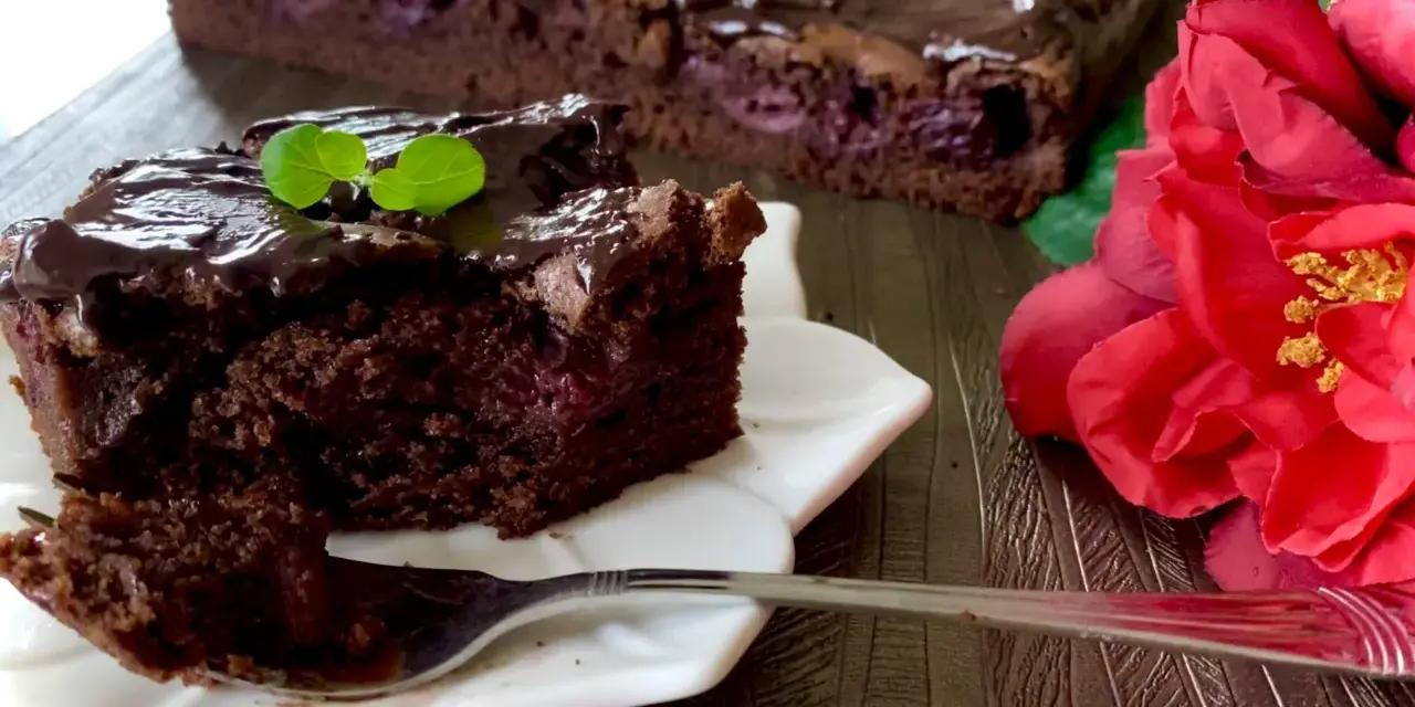 Pyszny brownie z wiśniami, polane czekoladą, z listkiem mięty. Obok czerwona róża.