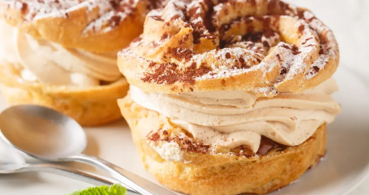 Dwa pyszne ciastka Paris-Brest z kremem kawowym, posypane kakao i cukrem pudrem, na talerzu ze srebrnymi łyżeczkami i miętą.