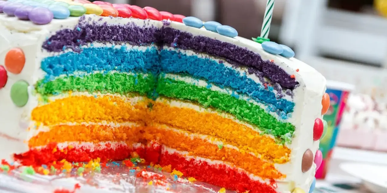 Kolorowy tort tęczowy z warstwami ciasta w kolorach tęczy, ozdobiony cukierkami i świeczką.