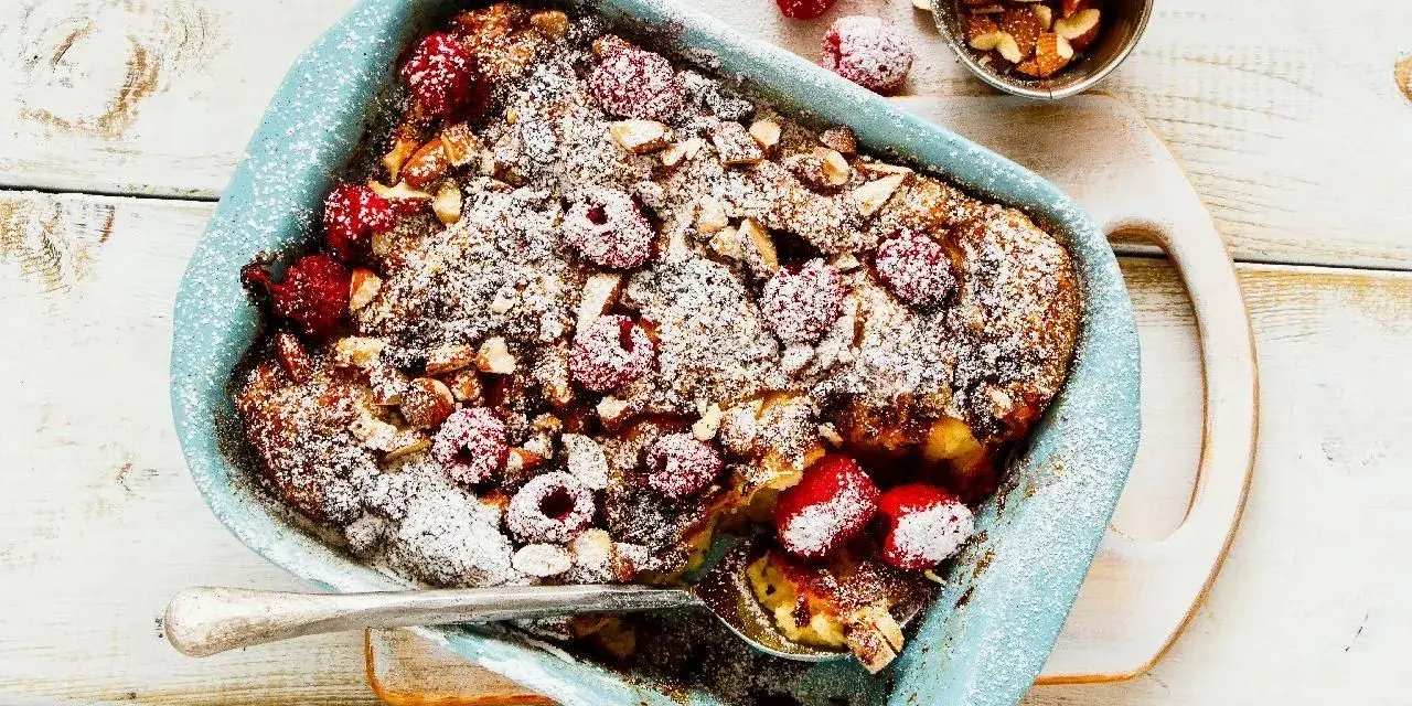 Pyszny pudding chlebowy z malinami i migdałami, posypany cukrem pudrem, w niebieskiej formie. Idealny deser.