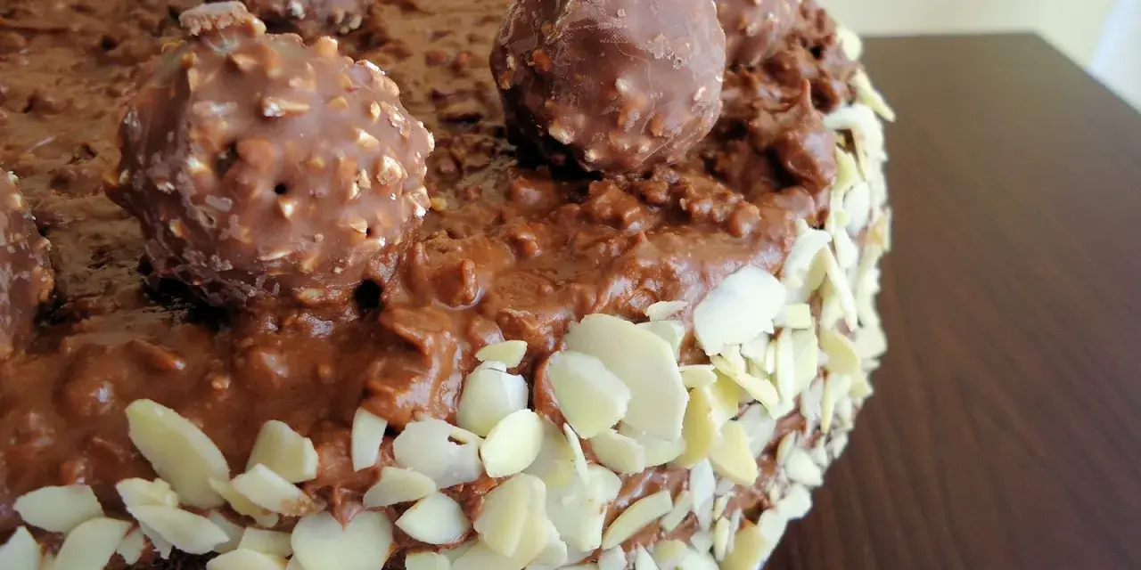 Pyszny tort Ferrero Rocher, oblany czekoladą i obsypany płatkami migdałów, z całymi czekoladkami na wierzchu.