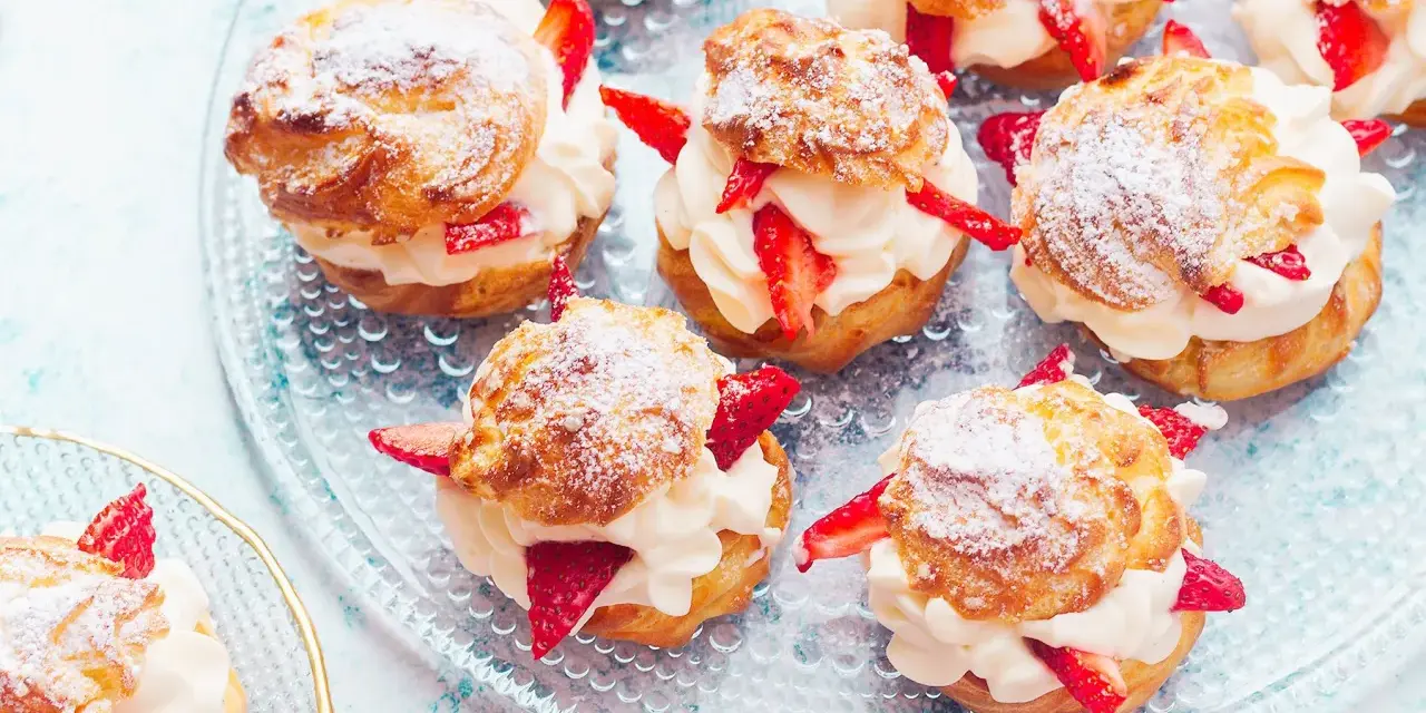 Pyszne ptysie z kremem patissiere i świeżymi truskawkami, posypane cukrem pudrem, prezentują się apetycznie na szklanej paterze.