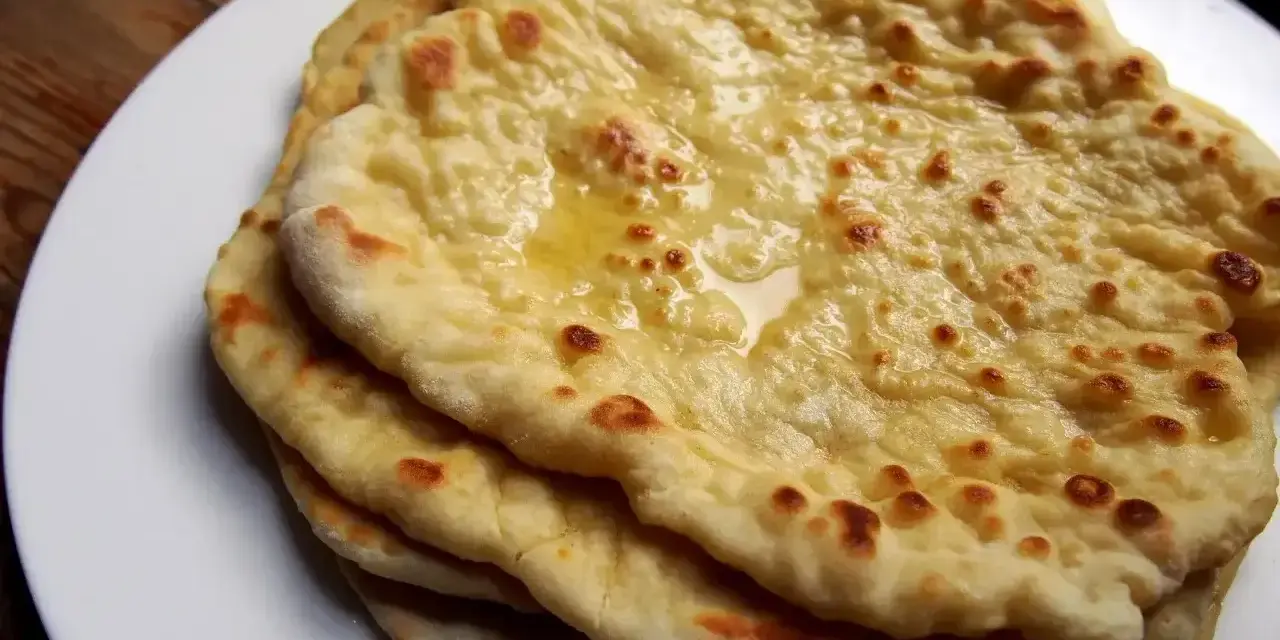 Stos świeżo upieczonych, lekko przypieczonych placków roti i naan, polanych roztopionym masłem, na białym talerzu.