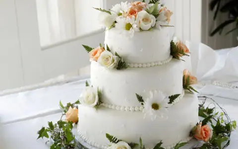 Elegancki tort weselny ozdobiony białymi i brzoskwiniowymi różami, margaretkami i zielonymi liśćmi. Piękne, jakie żywe kwiaty na tort!