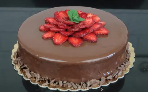 Lśniący tort czekoladowo truskawkowy, ozdobiony plasterkami truskawek i listkami mięty, otoczony czekoladowymi wiórkami.
