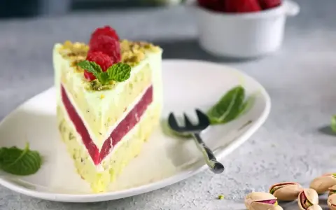 Pyszny tort pistacjowy z malinowym nadzieniem, udekorowany malinami i miętą. Obok leżą łuskane pistacje.
