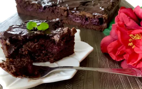 Pyszny brownie z wiśniami, polane czekoladą, z listkiem mięty. Obok czerwona róża.