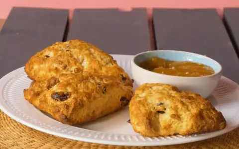 Złociste bułeczki scones z rodzynkami, podane na białym talerzu z dżemem. Idealne na popołudniową herbatkę.