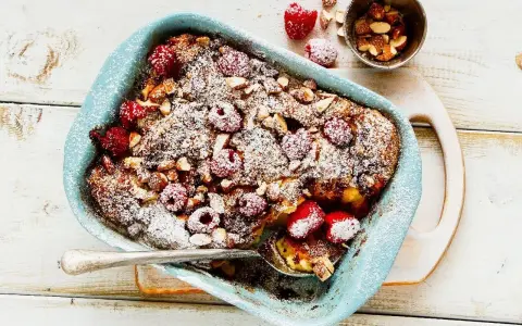 Pyszny pudding chlebowy z malinami i migdałami, posypany cukrem pudrem, w niebieskiej formie. Idealny deser.