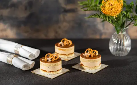 Trzy mini torty z kremem i polewą o smaku słonego karmelu, ozdobione precelkami i orzeszkami. Obok zwinięte serwetki.