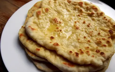 Stos świeżo upieczonych, lekko przypieczonych placków roti i naan, polanych roztopionym masłem, na białym talerzu.