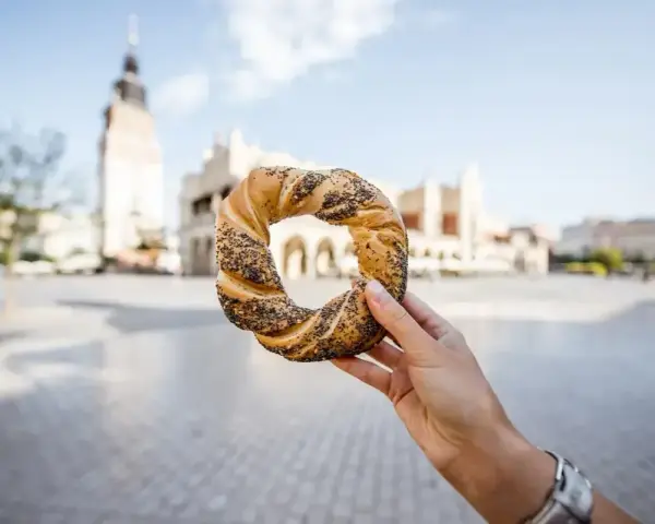 Ręka trzyma obwarzanka krakowskiego na tle Sukiennic i wieży Ratuszowej.