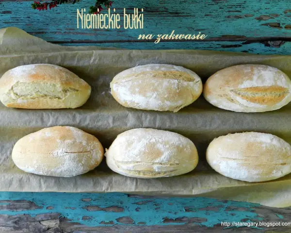 Sześć puszystych bułek na zakwasie, lekko oprószonych mąką, ułożonych na papierze do pieczenia.