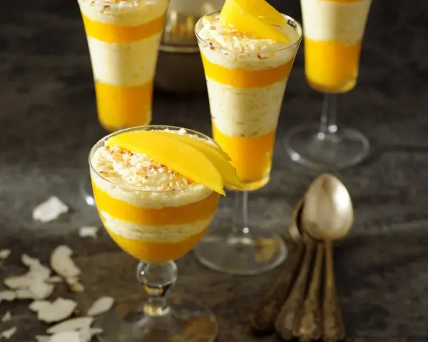 Warstwowy deser z mango, przełożony kremem kokosowym, ozdobiony plasterkiem mango i wiórkami kokosowymi.