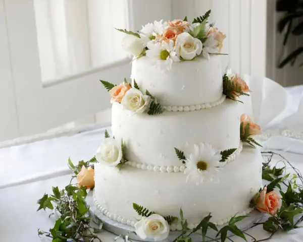 Elegancki tort weselny ozdobiony białymi i brzoskwiniowymi różami, margaretkami i zielonymi liśćmi. Piękne, jakie żywe kwiaty na tort!