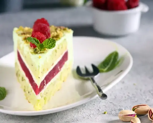 Pyszny tort pistacjowy z malinowym nadzieniem, udekorowany malinami i miętą. Obok leżą łuskane pistacje.