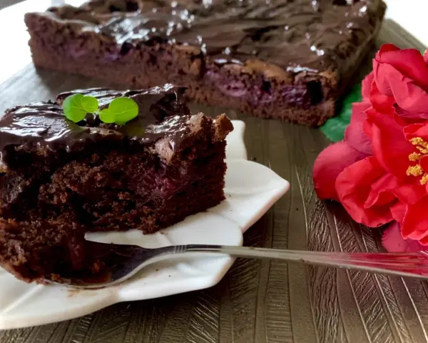 Pyszny brownie z wiśniami, polane czekoladą, z listkiem mięty. Obok czerwona róża.