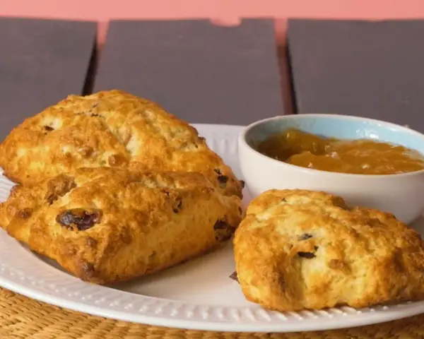 Złociste bułeczki scones z rodzynkami, podane na białym talerzu z dżemem. Idealne na popołudniową herbatkę.