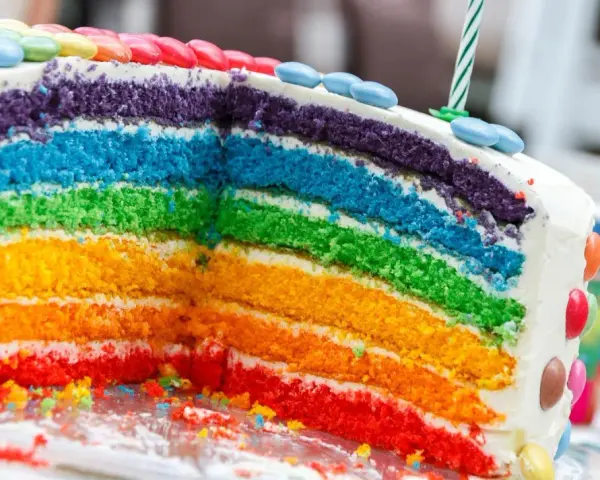 Kolorowy tort tęczowy z warstwami ciasta w kolorach tęczy, ozdobiony cukierkami i świeczką.