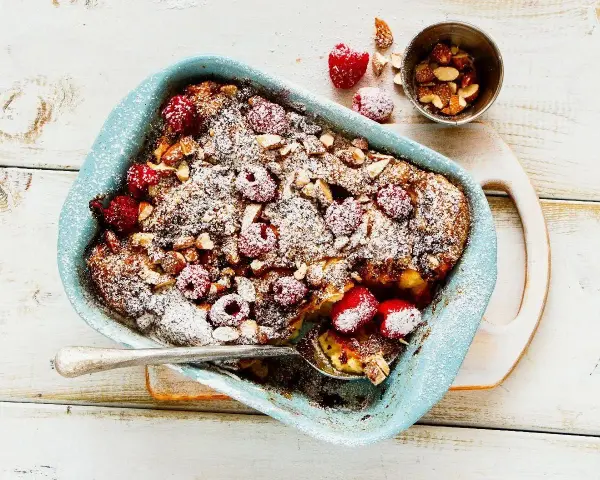 Pyszny pudding chlebowy z malinami i migdałami, posypany cukrem pudrem, w niebieskiej formie. Idealny deser.