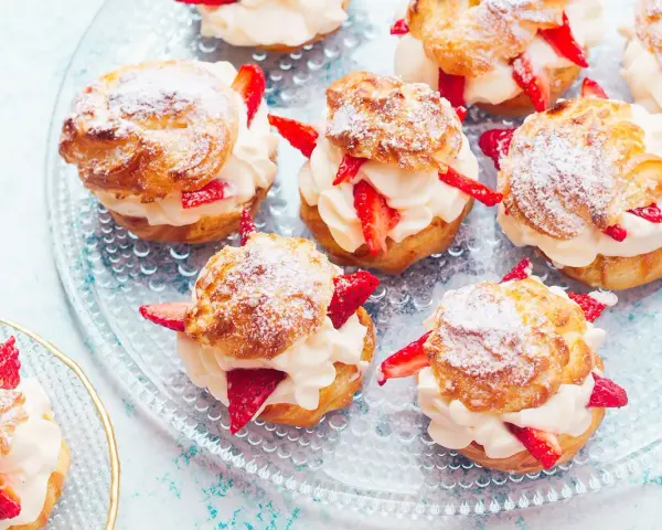 Pyszne ptysie z kremem patissiere i świeżymi truskawkami, posypane cukrem pudrem, prezentują się apetycznie na szklanej paterze.