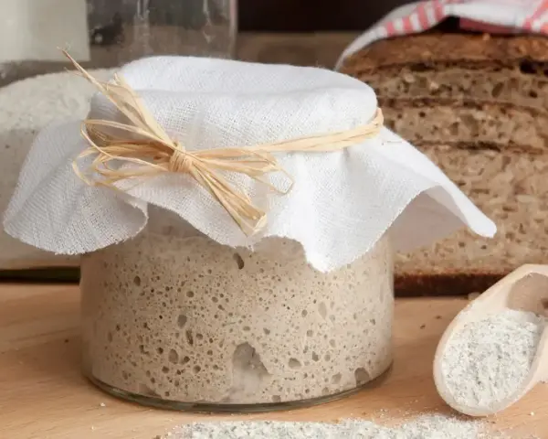 Słoik z zakwasem na chleb, obok kromki chleba i mąka w drewnianej łyżce. Nawet czerstwy chleb zasługuje na nowy początek.