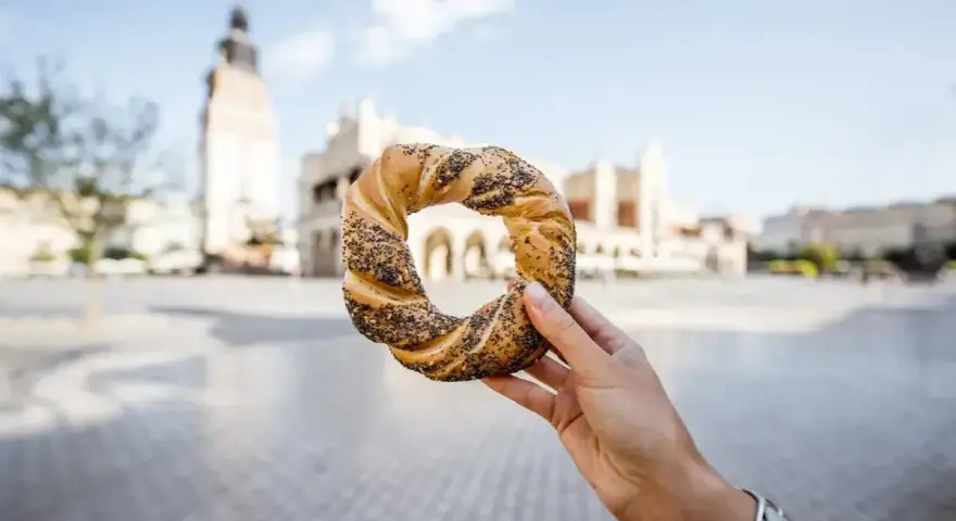 Ręka trzyma obwarzanka krakowskiego na tle Sukiennic i wieży Ratuszowej.