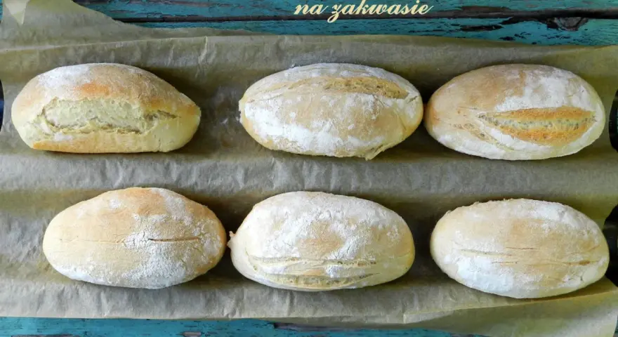 Sześć puszystych bułek na zakwasie, lekko oprószonych mąką, ułożonych na papierze do pieczenia.