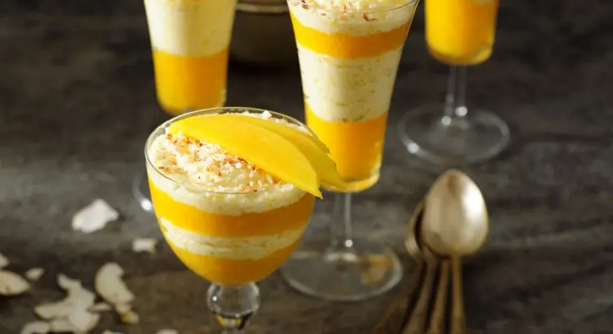 Warstwowy deser z mango, przełożony kremem kokosowym, ozdobiony plasterkiem mango i wiórkami kokosowymi.