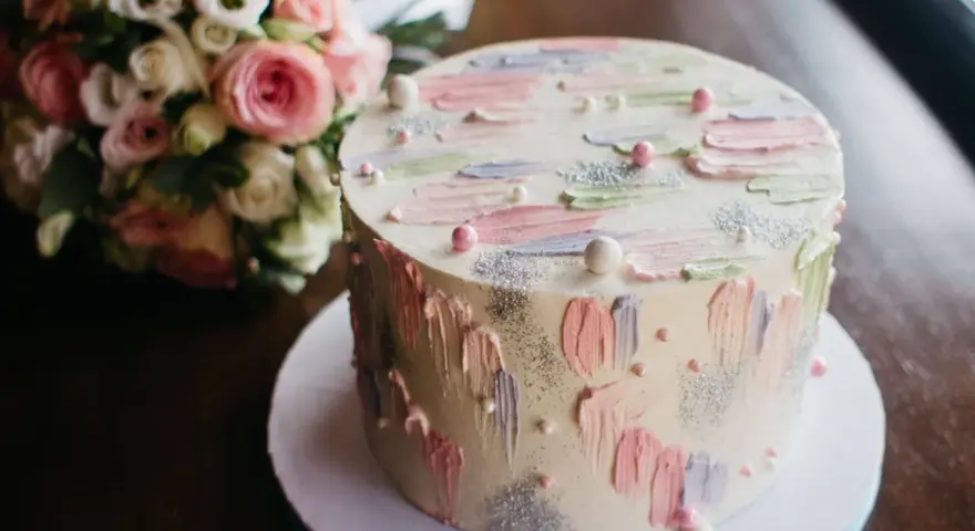 Oto tort na 20 osób, ważący około 2 kg, ozdobiony pastelowymi pociągnięciami pędzla i perełkami. Obok bukiet różowych kwiatów.
