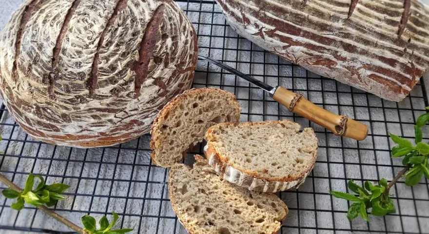Dwa bochenki chleba żytniego, jeden okrągły, drugi podłużny, leżą na kratce. Obok pokrojone kromki, nóż z drewnianą rączką i zielone gałązki.