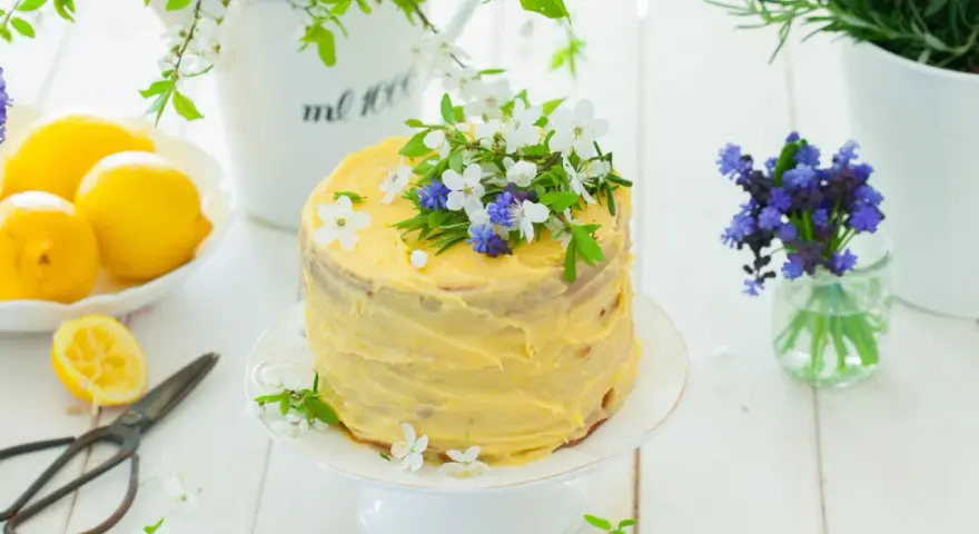 Słoneczny tort cytrynowy z kremem lemon curd, ozdobiony świeżymi kwiatami i ziołami. Obok leżą cytryny i stare nożyczki.