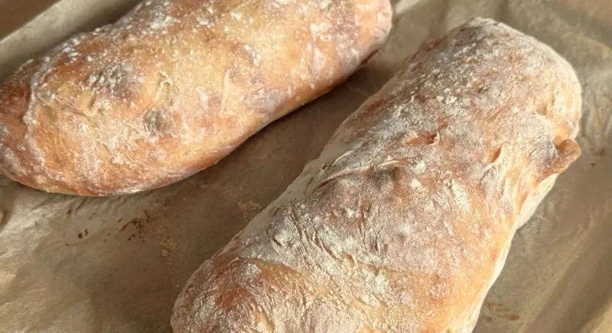 Dwa świeżo upieczone bochenki ciabatty, posypane mąką, leżą na papierze do pieczenia. Idealne do wypróbowania tego ciabatta przepis!