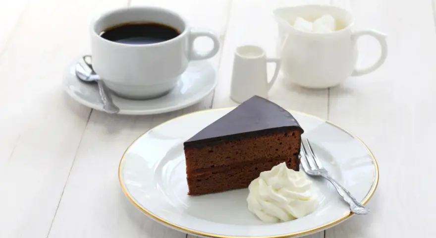 Pyszny tort Sachera z bitą śmietaną i kawą na białym drewnianym stole. Idealny deser na chwilę relaksu.