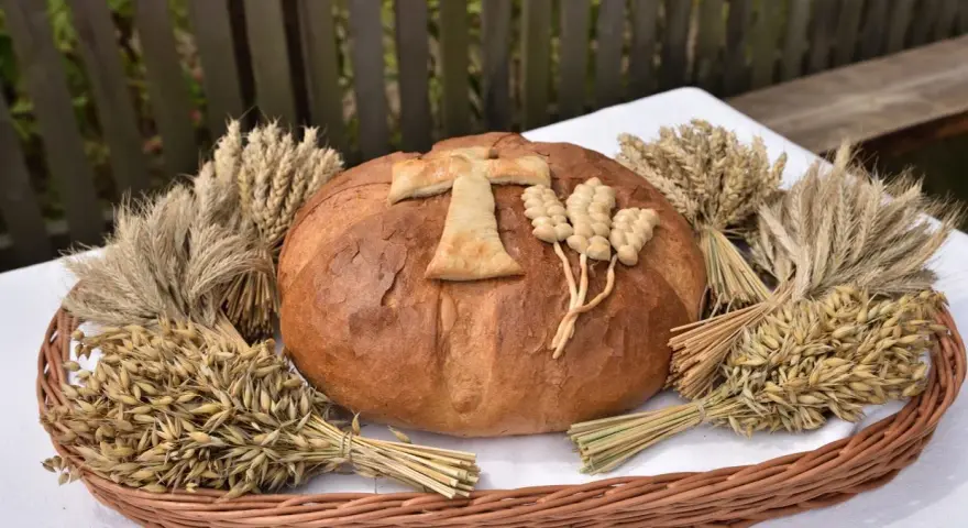 Chleb dożynkowy ozdobiony krzyżem i kłosami, otoczony snopkami zbóż. Inspiracja, jak ozdobić chleb dożynkowy.