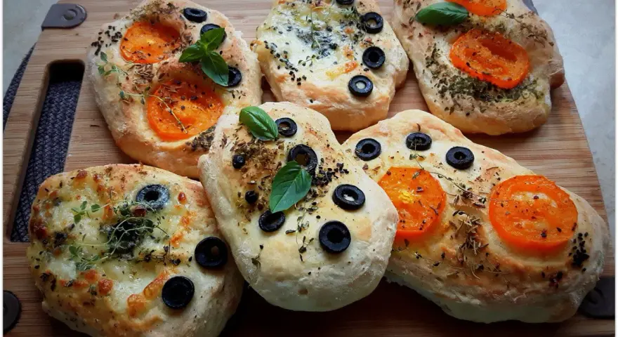Mini ciabatty z oliwkami, pomidorkami i ziołami, pięknie ułożone na desce.