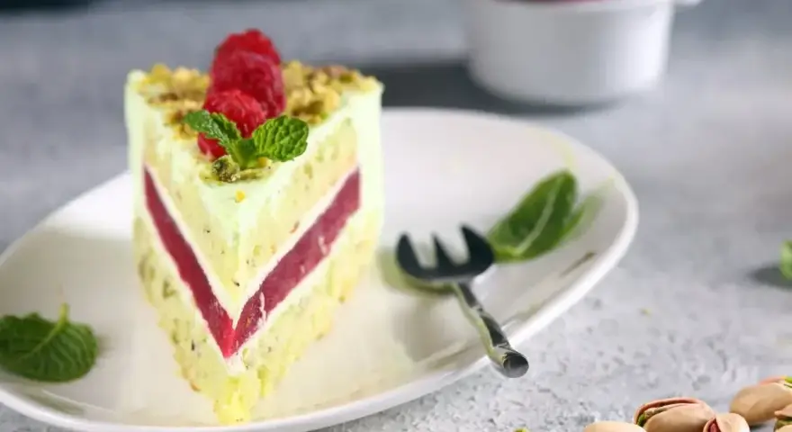 Pyszny tort pistacjowy z malinowym nadzieniem, udekorowany malinami i miętą. Obok leżą łuskane pistacje.