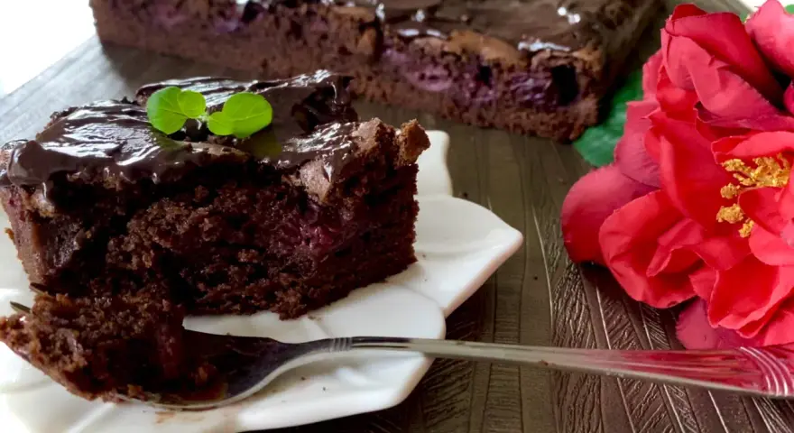 Pyszny brownie z wiśniami, polane czekoladą, z listkiem mięty. Obok czerwona róża.