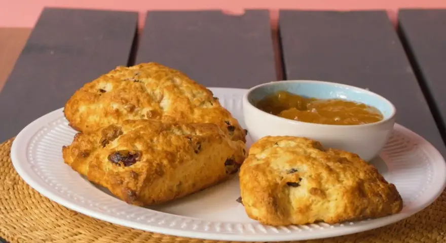 Złociste bułeczki scones z rodzynkami, podane na białym talerzu z dżemem. Idealne na popołudniową herbatkę.