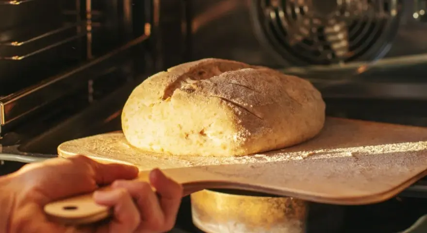 Ręka wsuwa drewnianą łopatą bochenek chleba do rozgrzanego piekarnika. To kulminacyjny moment wyrabiania ciasta na chleb.