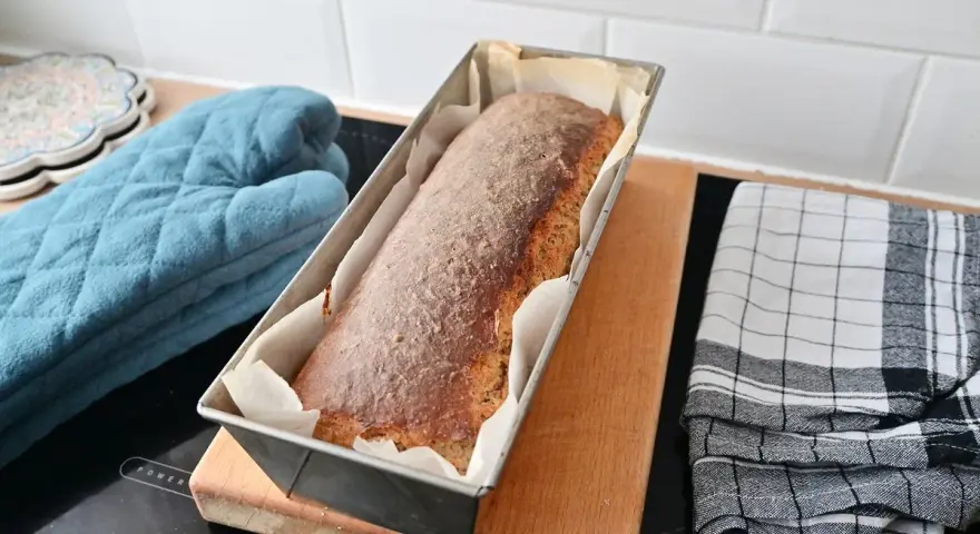 Świeżo upieczony chleb cebulowy na zakwasie w keksówce, gotowy do wyjęcia. Obok rękawica kuchenna i ściereczka.