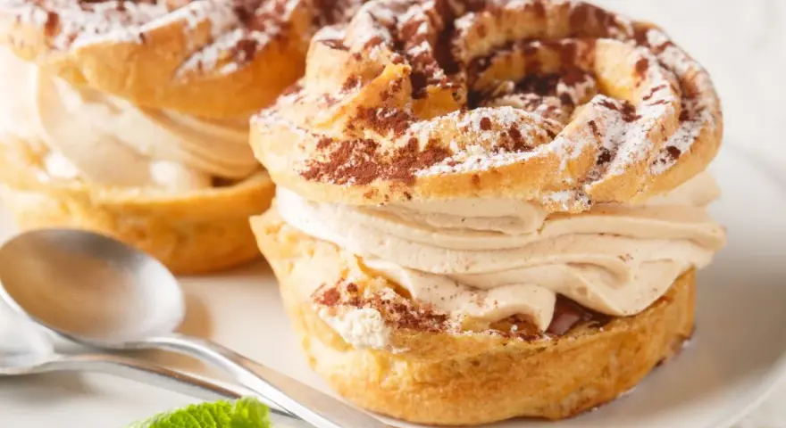 Dwa pyszne ciastka Paris-Brest z kremem kawowym, posypane kakao i cukrem pudrem, na talerzu ze srebrnymi łyżeczkami i miętą.