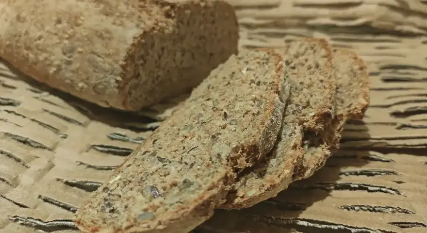 Chleb orkiszowy, pokrojony w kromki, z widocznymi ziarnami, leży na falistej tekturze.