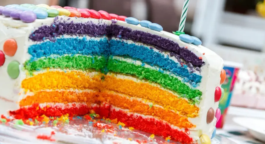 Kolorowy tort tęczowy z warstwami ciasta w kolorach tęczy, ozdobiony cukierkami i świeczką.