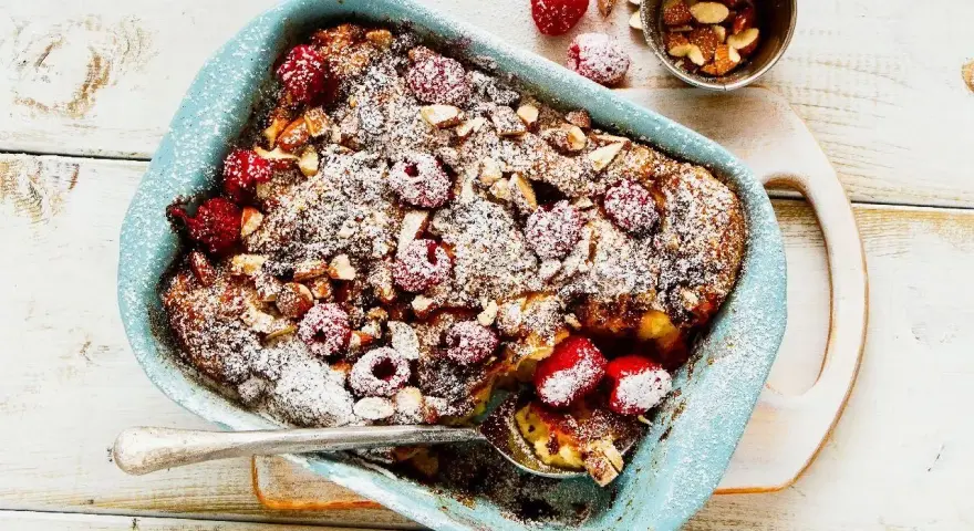 Pyszny pudding chlebowy z malinami i migdałami, posypany cukrem pudrem, w niebieskiej formie. Idealny deser.